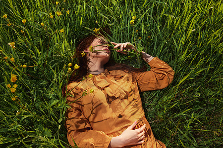 红色长围裙摄影照片_一个长着红色长发的平静女人躺在黄色花朵的绿色田野里，穿着橙色的连衣裙，闭着眼睛，用手抚摸着草叶，享受着平静和休养