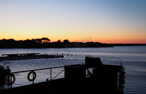 一轮太阳摄影照片_暮色笼罩着一个湖，太阳落在一个树木覆盖的岛屿后面，岛上有闪闪发光的橙色天空和一艘码头和旧桨船的轮廓
