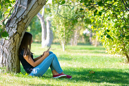 一个女孩坐在阳光明媚的公园的树下，看着手机屏幕