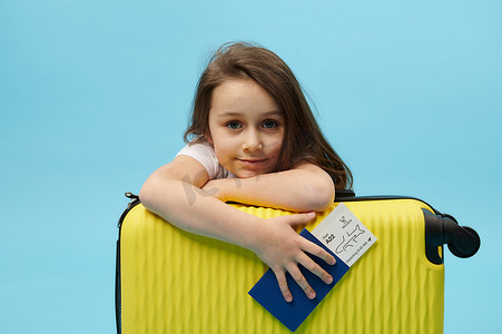快乐的旅行者孩子，可爱的 5 岁长发小女孩，拿着登机牌，看着黄色手提箱后面的相机，孤立的蓝色背景。