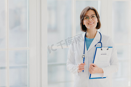 漂亮的微笑实习生拿着写有纸和笔的剪贴板，穿着白色制服，准备在自己的私人诊所看病人，站在室内白色背景下。
