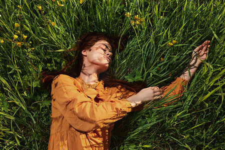 红色长围裙摄影照片_一个长着红色长发的平静女人躺在一片高高的草丛中，身着橙色连衣裙，闭着眼睛，在她面前伸展双臂