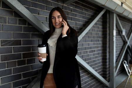 四十岁的女商人手里拿着咖啡，在一栋建筑的砖墙背景下用手机说话