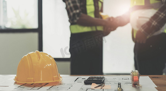 工作场所办公桌上的黄色安全帽与建筑工人团队握手问候启动计划在建筑工地办公中心的新项目合同、伙伴关系和承包商概念