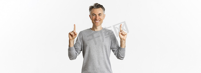 男士促销摄影照片_留着灰色短发的英俊成功男士的画像，高兴地笑着，用手指指着标志，展示促销优惠，站在白色背景上