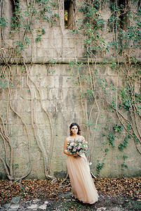 拿着花束的新娘站在常春藤缠绕的石头堡垒墙附近