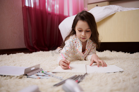人才发展摄影照片_穿着睡衣的可爱小女孩躺在卧室的地毯上，专心致志地用彩色铅笔画画