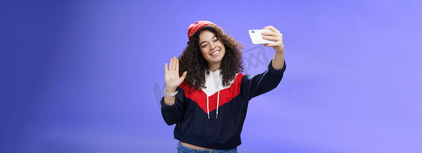 studio摄影照片_Studio shto 的时尚可爱女性视频博主，头发卷曲，头戴冬季红帽，在智能手机摄像头上微笑并挥手打招呼，录制视频，制作有趣的内容，在蓝墙上发布到网上