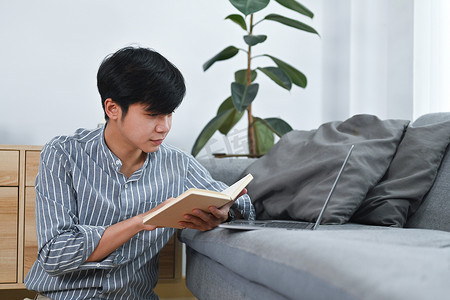 愉快的亚洲人坐在地板上看书，在家里度过闲暇时光。