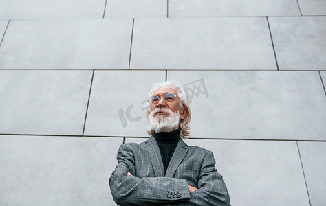 灰白的头发摄影照片_身着正装、头发和胡须灰白的高级商人在户外靠墙站立