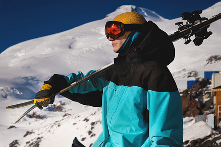 一幅戴着橙色滑雪护目镜的职业滑雪者的肖像站在雪山的背景下，肩上扛着滑雪板。