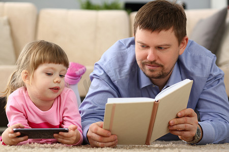 爸爸给女儿看书，女儿拿着智能手机躺在地上
