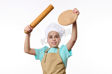 儿童拿着相机摄影照片_穿着厨师围裙和帽子的快乐青春期前儿童拿着木板和滚针，看着相机，白色背景