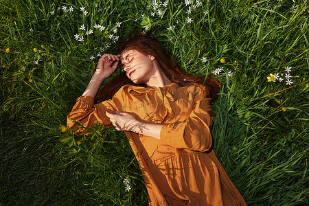 一个轻松的女人，躺在绿草地上休息，穿着橙色的长裙，闭着眼睛，脸上挂着愉快的微笑，正在休养