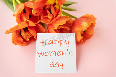 打卡美陈摄影照片_一束美丽的橙色牡丹郁金香，中间有一张白卡，粉红色背景上写着妇女节快乐。