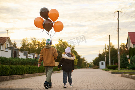 小学生、男孩和女孩的后视图，带着橙色黑色气球在万圣节的乡村街道上行走