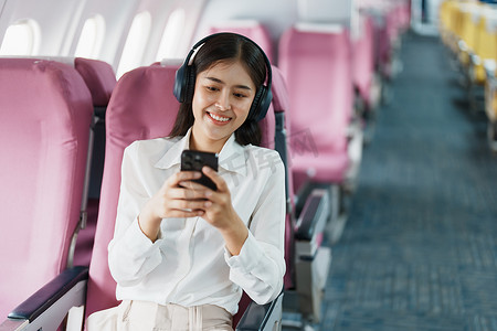 年轻的亚洲美女乘客坐在商务舱豪华飞机上，同时戴着无线耳机，在飞行期间使用智能手机放松