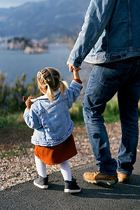 爸爸拿摄影照片_爸爸牵着一个拿着毛绒玩具的小女孩的手，站在路上以大海、高山和圣斯特凡岛为背景。