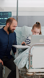 父亲坐在生病的女儿身边，用笔记本电脑玩卡通在线视频游戏