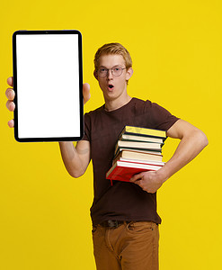 融合教育摄影照片_惊讶的学生拿着书和平板电脑，上面有空白的白色屏幕，用于黄色背景的复制空间，展示了现代教育的概念以及技术在学习和学习中的融合。