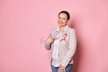 积极的多民族女性，站在粉红色背景上，用手指指着夹克上的粉红色缎带