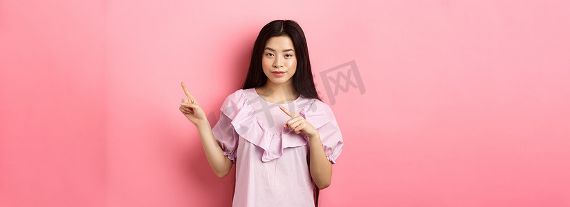 厚颜无耻的亚洲女孩展示有趣的广告，用手指指着标志，狡猾地微笑，暗示很划算，粉红色背景