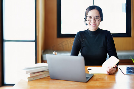 一位戴着眼镜、使用电脑笔记本电脑、耳机并使用笔记本电脑通过木制图书馆桌上的视频会议在线学习的亚洲少女肖像