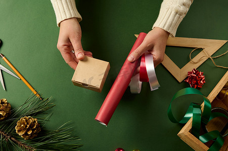 女人拿着小纸箱、红色包装纸和装饰闪亮的丝带，用于在绿色表面包装礼物