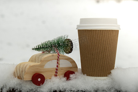 木制汽车载着圣诞树，纸杯在雪地上模拟咖啡或热巧克力。