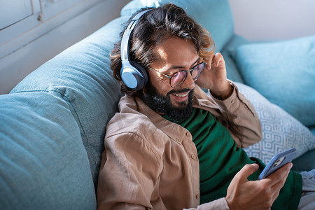 戴着耳机微笑的年轻人坐在家里的沙发上听智能手机播放的音乐