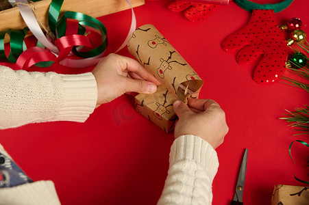 圣诞快乐小鹿摄影照片_特写女人用红色背景上带有鹿图案的礼品纸包裹着一份小礼物。