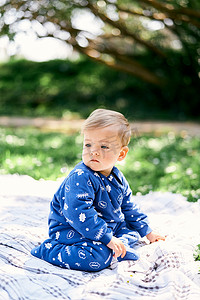 身穿蓝色工装裤的小孩跪在绿色草坪上的方格毯子上，头转向
