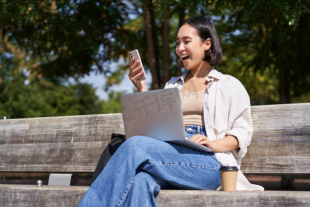 兴奋的亚洲年轻女孩看着她的智能手机，在阳光明媚的公园里坐在户外的笔记本电脑上