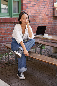 时尚亚洲女孩独自坐在咖啡馆里，带着笔记本电脑、工作或学习、在电脑上浏览的垂直照片