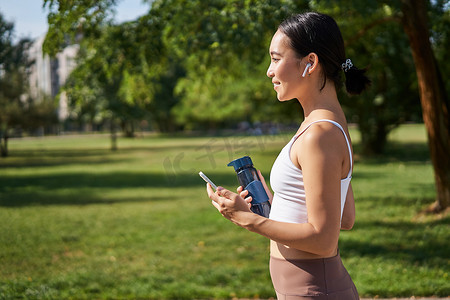 活跃的亚洲女孩，穿着健身服，在公园锻炼，穿着带智能手机和水瓶的运动服走路