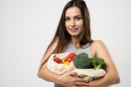 穿着灰色连衣裙的快乐微笑的女人拿着可重复使用的网袋，里面装着白色背景的水果和蔬菜。