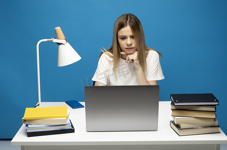 t恤女摄影照片_年轻成功的秘书雇员女商人的工作室肖像穿着白色 T 恤坐在白色办公桌前工作，笔记本电脑在线浏览互联网隔离在蓝色背景。