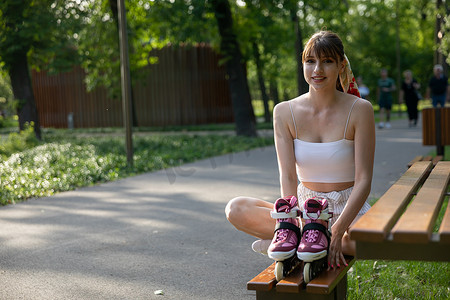 一个年轻的微笑女孩坐在城市公园的长椅上，准备好旱冰鞋。