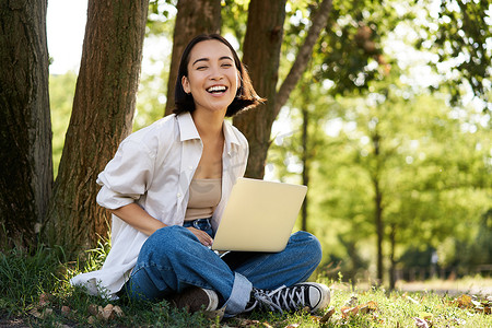 亚洲女孩带着笔记本电脑的肖像，在阳光明媚的夏日坐在公园的树旁，用电脑进行远程、电子学习