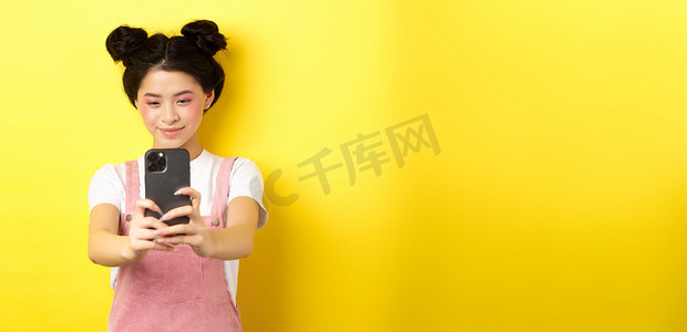 时尚的亚洲女孩在智能手机上拍照，用手机拍视频，微笑着，站在黄色背景上