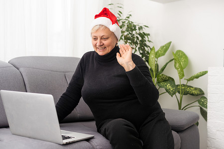 圣诞节老人摄影照片_圣诞节时，戴着圣诞帽的微笑高级女性在家里用笔记本电脑打字，与家人或朋友取得联系。