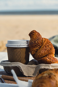 咖啡原料摄影照片_美味的新鲜羊角面包和沙滩海岸背景纸杯中的咖啡。