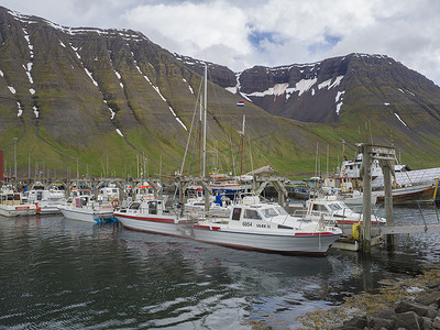 冰岛、西峡湾、伊萨菲厄泽，2018 年 6 月 25 日：伊萨菲厄泽港的夏日景色，有渔船和美丽的绿色雪山和悬崖、云彩和蓝天背景