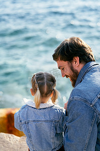 坐在爸爸的背上摄影照片_微笑的爸爸坐在海面上一块石头上的一个小女孩旁边。