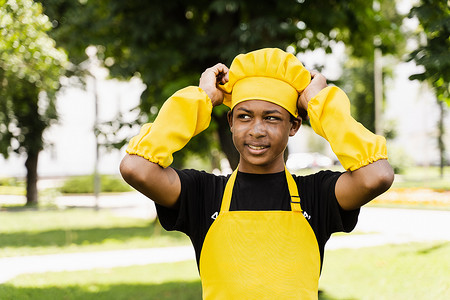 身着厨师帽和黄色围裙制服的非洲黑人少年厨师摸着他的厨师帽。