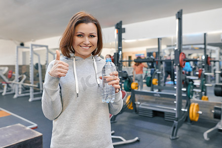 成熟的女人在健身房里拿着一瓶水，并表现出 ok 的手势。