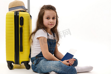 旅行者护照摄影照片_身穿蓝色牛仔服、登机牌和黄色手提箱的小女孩旅行者，在白色背景中突显