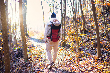 婴儿背包摄影照片_运动型父亲背着他的小儿子穿着冬季连身衣和帽子，背着背包在秋天的森林里徒步旅行。