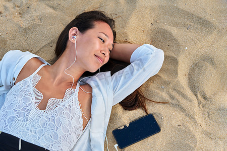 戴着耳机、在沙滩上用智能手机放松的年轻女性，俯视