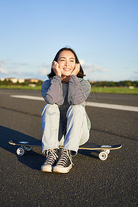 亚洲女滑手的垂直镜头，坐在她的滑板上微笑，享受阳光明媚的日子，在户外空荡荡的路上在长板上巡航